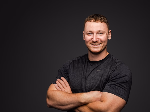Ein lächelnder Mann mit verschränkten Armen, trägt ein schwarzes Sportshirt, vor dunklem Hintergrund.